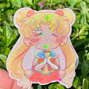 Può includere: Un adesivo olografico raffigurante un personaggio dei cartoni animati con lunghi capelli biondi, un'uniforme da marinaio blu e una gonna rossa. Il personaggio è seduto con le mani giunte. L'adesivo ha un bordo rosa ed è decorato con stelle e diamanti.