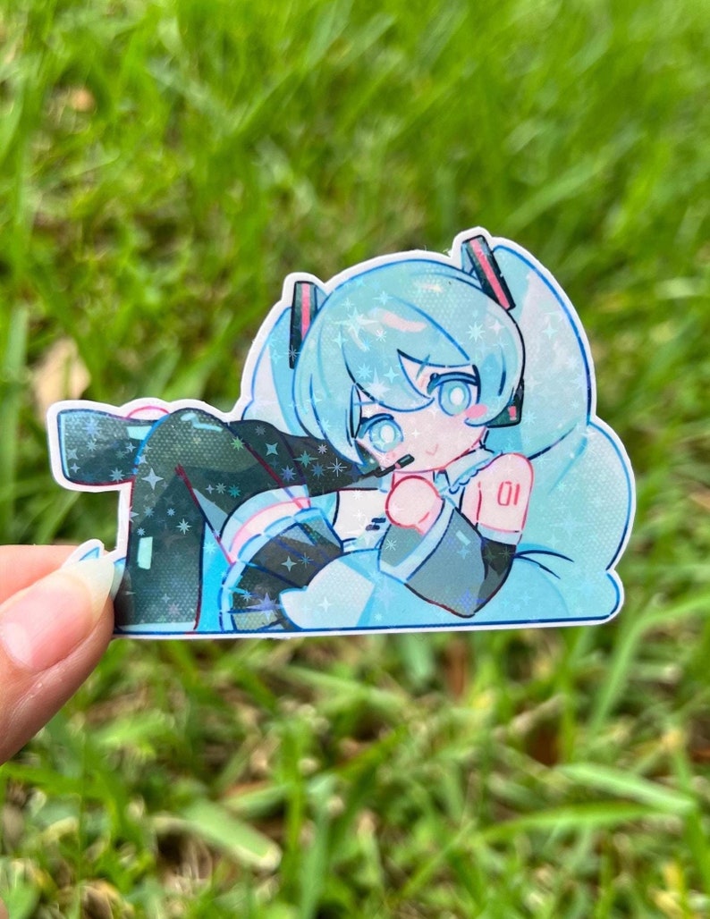May include: A holographic sticker featuring a stylized anime character with light blue hair and eyes. The character is wearing a black and teal outfit with the number "01" visible. The sticker has a sparkly, glittery effect.
