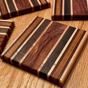 Op de afbeelding: Vier vierkante houten onderzetters met een gestreept patroon in tinten bruin, zwart en beige. De onderzetters zijn gemaakt van verschillende soorten hout, wat een unieke en rustieke uitstraling creëert.