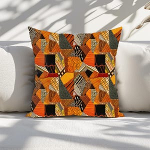 May include: Square throw pillow with a geometric pattern in shades of orange, black, and gray. The design features a patchwork of triangles and other shapes with various textures and patterns. The pillow is resting on a white surface.