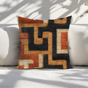 May include: Square decorative pillow with a geometric pattern in shades of black, orange, tan, and cream. The pillow is placed on a white surface, with two white pillows on either side. The design features a maze-like pattern.