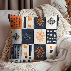 May include: Square throw pillow with a patchwork design. The pillow features a repeating pattern of squares in navy blue, orange, and cream. The squares contain various geometric and sun-like shapes. The pillow is resting on a beige couch with a cream-colored blanket.
