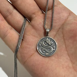 May include: A silver-colored pendant necklace featuring a detailed dragon design within a circular frame. The dragon is intricately sculpted, and the chain has a textured, woven appearance. The pendant hangs from a delicate chain.