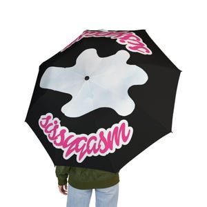 May include: A black umbrella featuring a large, central white cloud-like shape and the text "Sissygasm" printed in a bold, bright pink font with a white outline. The design is repeated in a circular pattern across the canopy of the umbrella.
