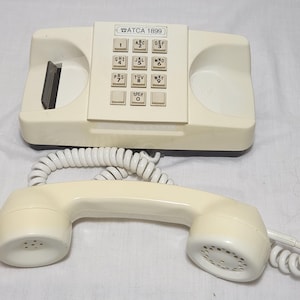 May include: Cream-colored vintage telephone with a handset and a base featuring a keypad with numbers and letters. The phone has a coiled cord and the text "ATCA 1899" is visible.