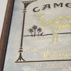 Puede incluir: Un letrero de espejo enmarcado con una ilustraci&oacute;n dorada de un camello, palmeras y una pir&aacute;mide. El texto dice R.J. Reynolds Tobacco Co., Camel, I'd walk a mile for a Camel y 1875 Centennial. El letrero est&aacute; dentro de un marco de madera marr&oacute;n.