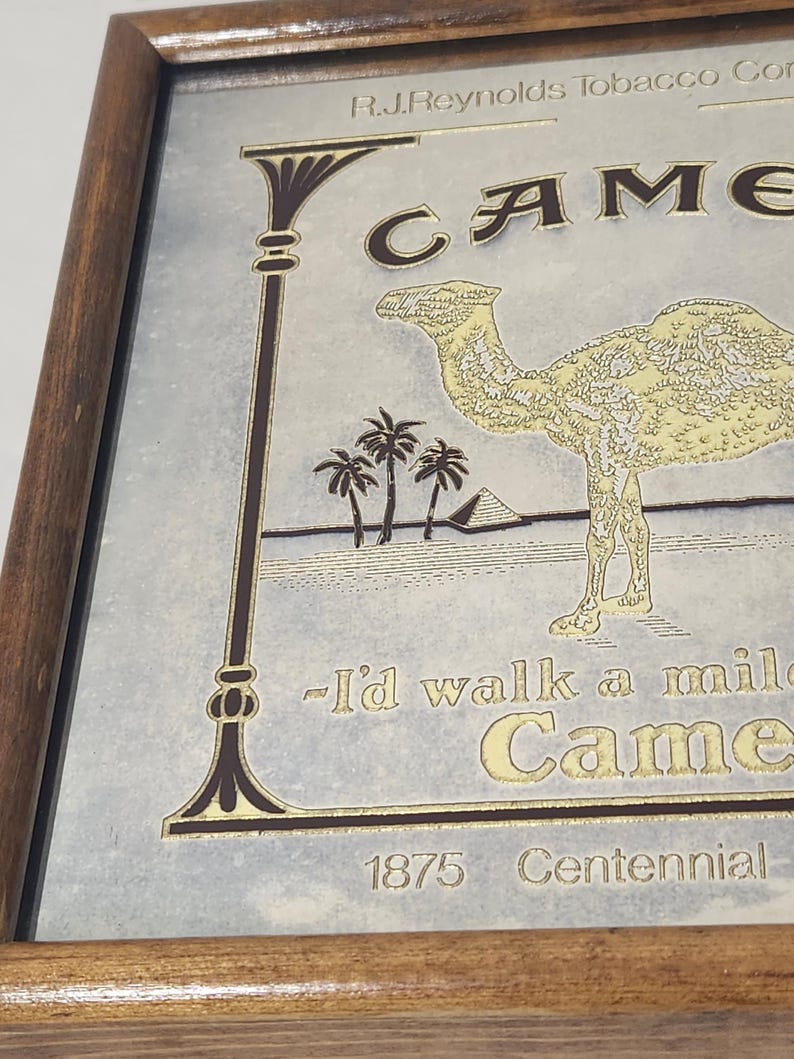 Puede incluir: Un letrero de espejo enmarcado con una ilustraci&oacute;n dorada de un camello, palmeras y una pir&aacute;mide. El texto dice R.J. Reynolds Tobacco Co., Camel, I'd walk a mile for a Camel y 1875 Centennial. El letrero est&aacute; dentro de un marco de madera marr&oacute;n.