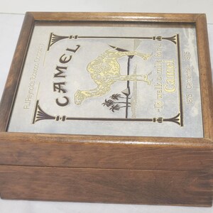 Puede incluir: Una caja cuadrada de madera con tapa abatible y panel superior met&aacute;lico. El panel muestra un gr&aacute;fico de camello dorado, palmeras y pir&aacute;mides. El texto incluye "CAMEL", "R.J. Reynolds Tobacco Company" y "I'd walk a mile for a Camel 1875-1975".
