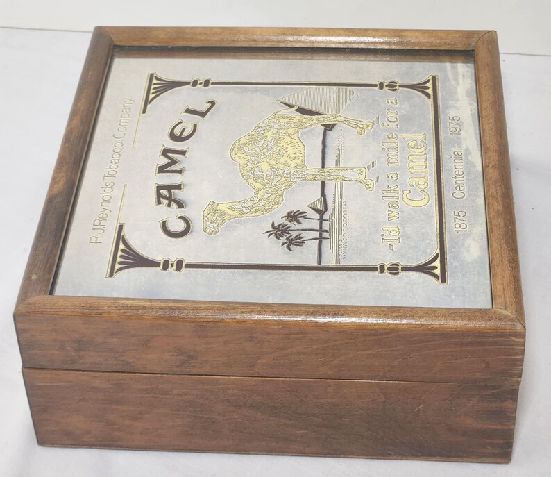 Puede incluir: Una caja cuadrada de madera con tapa abatible y panel superior met&aacute;lico. El panel muestra un gr&aacute;fico de camello dorado, palmeras y pir&aacute;mides. El texto incluye "CAMEL", "R.J. Reynolds Tobacco Company" y "I'd walk a mile for a Camel 1875-1975".