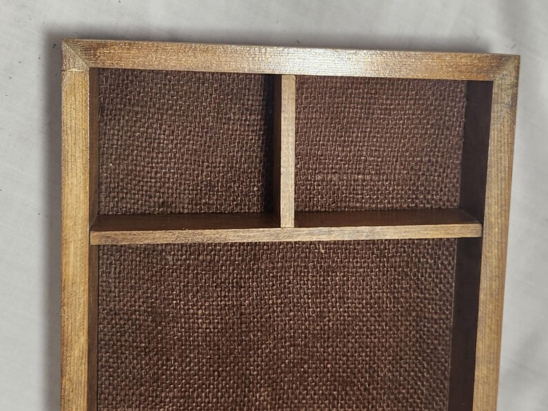 Puede incluir: Una bandeja rectangular de madera con acabado marr&oacute;n medio, que presenta un forro de tela texturizada en color marr&oacute;n oscuro. El interior est&aacute; dividido en tres secciones distintas por finos tabiques de madera, creando un &aacute;rea grande y dos compartimentos.