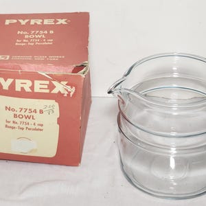 May include: A clear Pyrex bowl with a pouring spout, next to a red and white box. The box is labeled "PYREX" and "No. 7754 B BOWL". The bowl is made of clear glass and has a rounded shape. The box is slightly damaged.