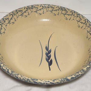 May include: A round, cream-colored ceramic bowl with a blue wheat stalk design in the center. The rim features a blue, textured pattern. This vintage-style bowl is suitable for serving food.