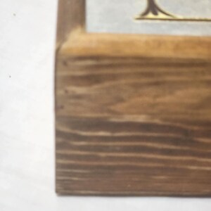 Puede incluir: Un marco de madera con acabado de veta marr&oacute;n claro que muestra una vista parcial de un inserto de color claro. El inserto presenta un elemento gr&aacute;fico decorativo marr&oacute;n oscuro con una base curva y una peque&ntilde;a parte de texto visible.