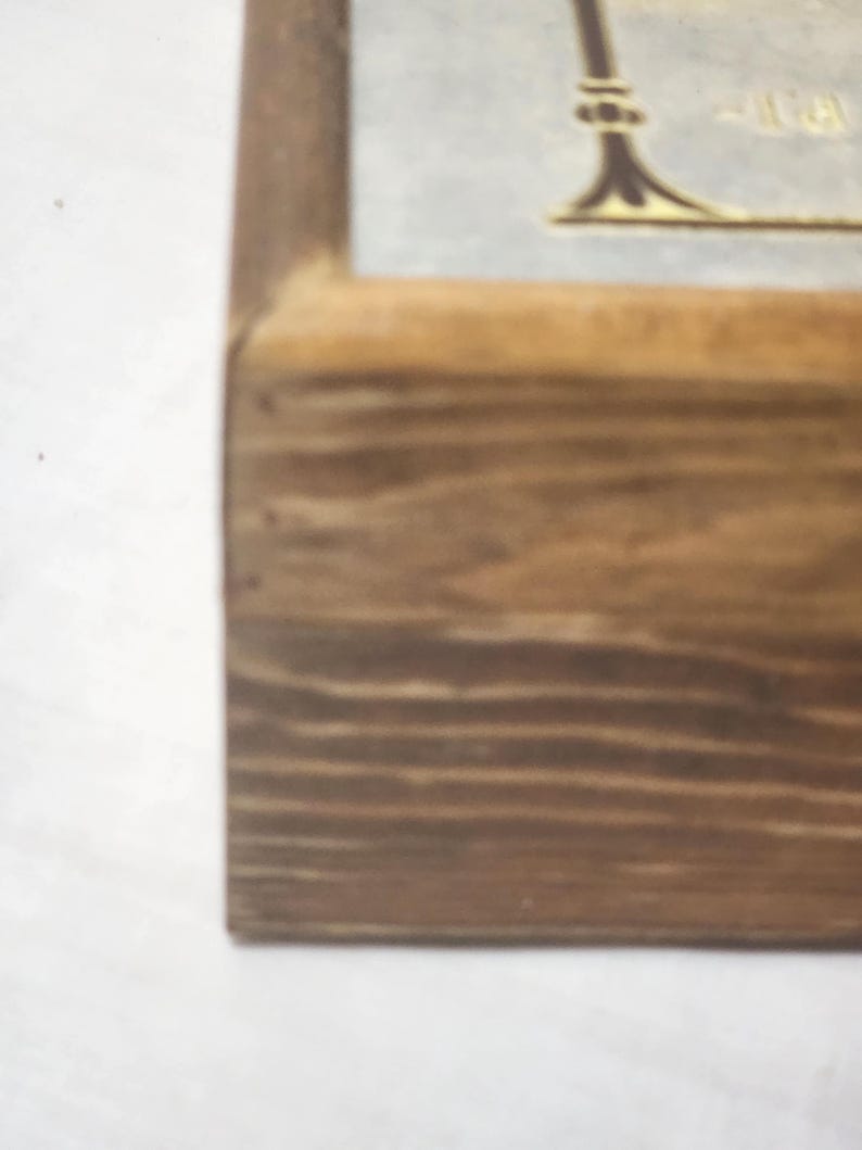 Puede incluir: Un marco de madera con acabado de veta marr&oacute;n claro que muestra una vista parcial de un inserto de color claro. El inserto presenta un elemento gr&aacute;fico decorativo marr&oacute;n oscuro con una base curva y una peque&ntilde;a parte de texto visible.
