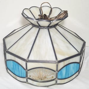 May include: A stained glass lamp shade with a scalloped top and an octagonal base. The shade features panels of white marbled glass accented with curved blue glass sections and a dried floral arrangement encased within one of the glass panels.