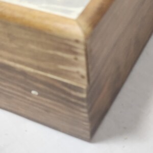 Puede incluir: Una caja rectangular de madera con un acabado de veta de madera marr&oacute;n natural. La tapa presenta un panel insertado liso de color claro con un dise&ntilde;o gr&aacute;fico en tono dorado visible en el borde superior. La caja tiene una estructura con bisagras.