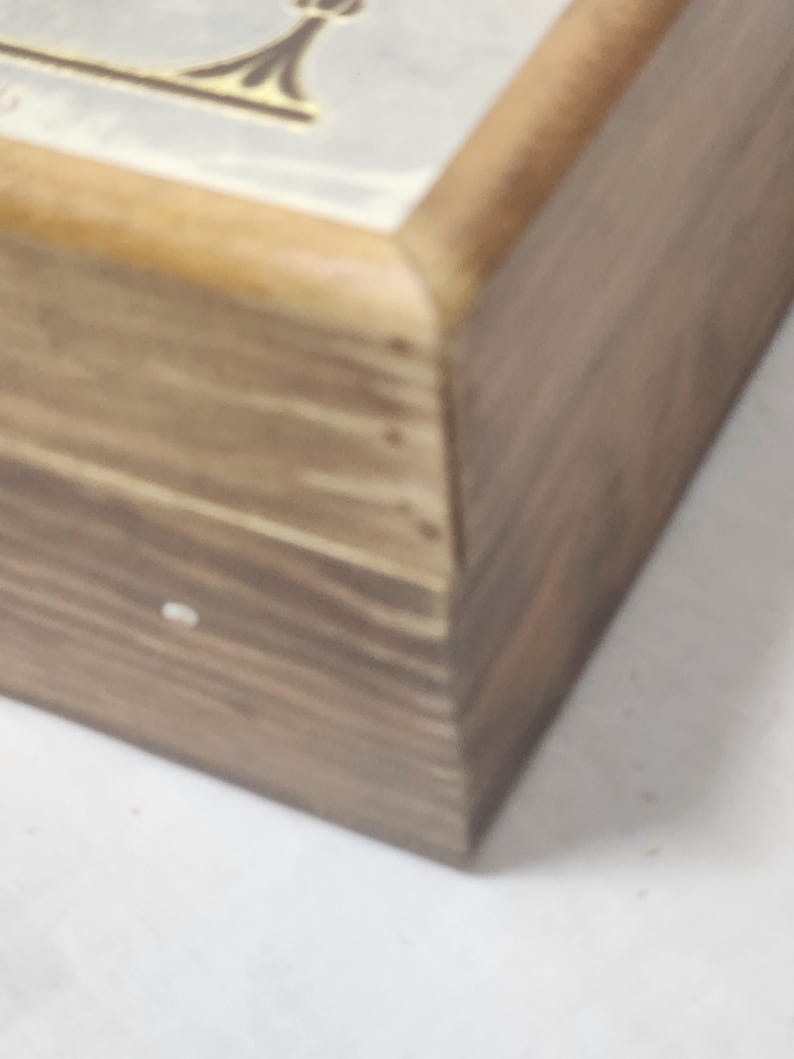 Puede incluir: Una caja rectangular de madera con un acabado de veta de madera marr&oacute;n natural. La tapa presenta un panel insertado liso de color claro con un dise&ntilde;o gr&aacute;fico en tono dorado visible en el borde superior. La caja tiene una estructura con bisagras.