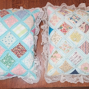 May include: Two square throw pillows featuring a cathedral window quilt pattern with various floral and geometric fabric patches in pastel and bright colors, separated by light blue fabric strips and finished with a white lace ruffle border.