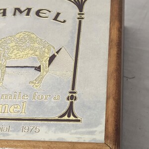 Puede incluir: Una caja de madera con un gr&aacute;fico de la marca Camel que muestra una ilustraci&oacute;n de un camello y una pir&aacute;mide. El texto dice R.J. Reynolds Tobacco Company, CAMEL, Smile for a Camel y 1975. El dise&ntilde;o est&aacute; impreso en dorado y marr&oacute;n oscuro.
