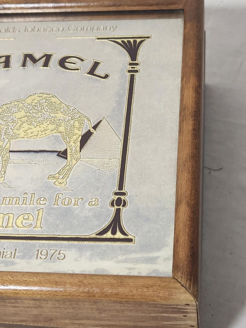 Puede incluir: Una caja de madera con un gr&aacute;fico de la marca Camel que muestra una ilustraci&oacute;n de un camello y una pir&aacute;mide. El texto dice R.J. Reynolds Tobacco Company, CAMEL, Smile for a Camel y 1975. El dise&ntilde;o est&aacute; impreso en dorado y marr&oacute;n oscuro.