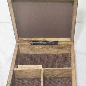 Puede incluir: Una caja cuadrada de madera con tapa abatible, con un acabado de veta de madera marr&oacute;n claro natural. El interior est&aacute; dividido en dos compartimentos con una base texturizada de color marr&oacute;n oscuro. Se ve una peque&ntilde;a etiqueta de papel.