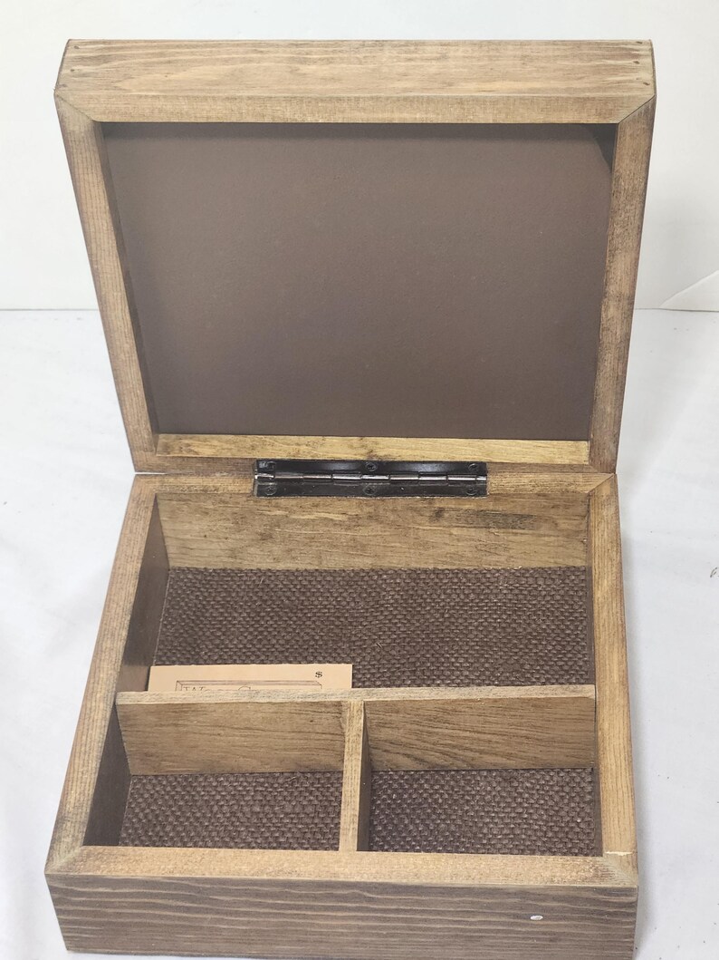 Puede incluir: Una caja cuadrada de madera con tapa abatible, con un acabado de veta de madera marr&oacute;n claro natural. El interior est&aacute; dividido en dos compartimentos con una base texturizada de color marr&oacute;n oscuro. Se ve una peque&ntilde;a etiqueta de papel.
