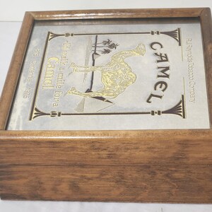 Puede incluir: Una caja cuadrada de madera con tapa abatible que presenta una inserci&oacute;n met&aacute;lica. La inserci&oacute;n muestra una ilustraci&oacute;n de un camello, pir&aacute;mides, palmeras y el texto: Camel, I'd walk a mile for a Camel, R.J. Reynolds Tobacco Company, 1875 Centennial 1975.