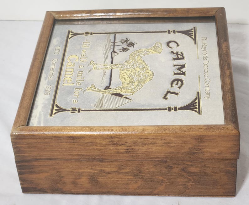 Puede incluir: Una caja cuadrada de madera con tapa abatible que presenta una inserci&oacute;n met&aacute;lica. La inserci&oacute;n muestra una ilustraci&oacute;n de un camello, pir&aacute;mides, palmeras y el texto: Camel, I'd walk a mile for a Camel, R.J. Reynolds Tobacco Company, 1875 Centennial 1975.