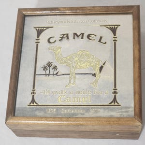 Puede incluir: Una caja de madera cuadrada con un gr&aacute;fico conmemorativo de cigarrillos Camel en la tapa. El dise&ntilde;o incluye un camello dorado, palmeras, pir&aacute;mides y el texto R.J. Reynolds Tobacco Company, Camel, I'd walk a mile for a Camel, 1875 Centennial 1975.