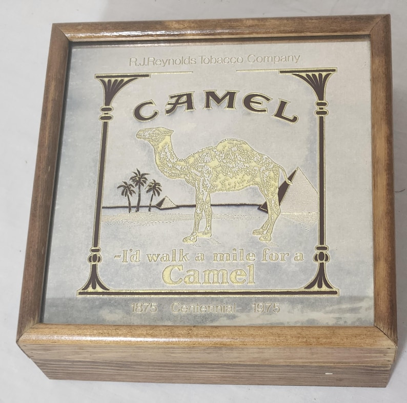 Puede incluir: Una caja de madera cuadrada con un gr&aacute;fico conmemorativo de cigarrillos Camel en la tapa. El dise&ntilde;o incluye un camello dorado, palmeras, pir&aacute;mides y el texto R.J. Reynolds Tobacco Company, Camel, I'd walk a mile for a Camel, 1875 Centennial 1975.
