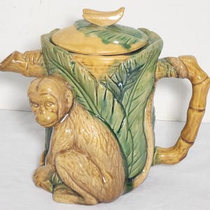 May include: A ceramic teapot with a monkey and banana design. The teapot features a monkey figure on the front, with a bamboo-style handle and spout. The lid is topped with a banana. The colors include tan, green, and brown.