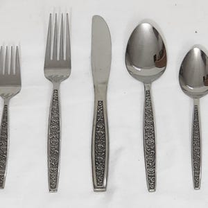 May include: A stainless steel flatware set featuring a fork, knife, and two spoons. The handles have a floral design. The set includes a dinner fork, a salad fork, a knife, a tablespoon, and a teaspoon.