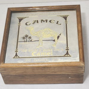 Puede incluir: Una caja cuadrada de madera con tapa de cristal que presenta un anuncio de cigarrillos Camel. El gr&aacute;fico incluye un camello, pir&aacute;mides, palmeras y el texto R.J. Reynolds Tobacco Company, Camel, I'd walk a mile for a Camel y 1875 Centennial 1975.