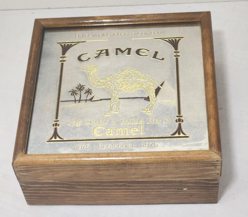 Puede incluir: Una caja cuadrada de madera con tapa de cristal que presenta un anuncio de cigarrillos Camel. El gr&aacute;fico incluye un camello, pir&aacute;mides, palmeras y el texto R.J. Reynolds Tobacco Company, Camel, I'd walk a mile for a Camel y 1875 Centennial 1975.
