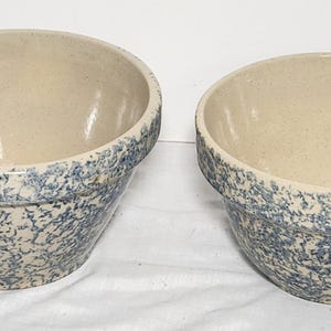 May include: Two stoneware mixing bowls. The bowls have a light beige interior and a blue speckled exterior. The rims of the bowls are also blue speckled. The bowls are sitting on a white surface.