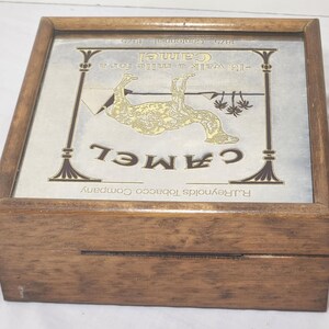 Puede incluir: Una caja cuadrada de madera con tapa abatible y un inserto de vidrio. El inserto muestra un anuncio de cigarrillos Camel con una ilustraci&oacute;n de un camello, el texto "I'd walk a mile for a Camel", "1875 Centennial 1975" y "R.J. Reynolds Tobacco Company".