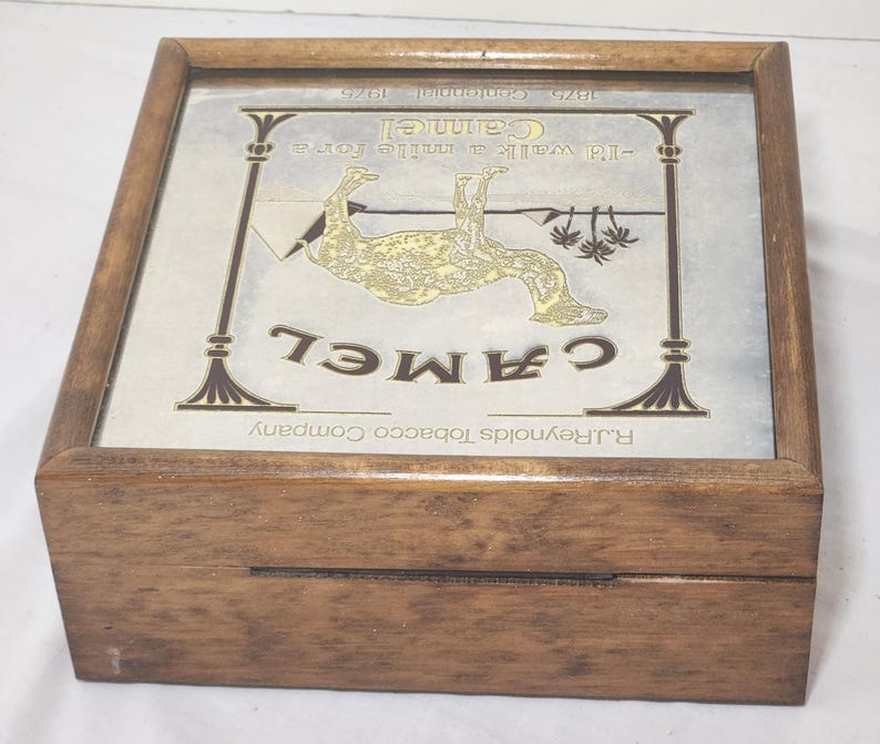 Puede incluir: Una caja cuadrada de madera con tapa abatible y un inserto de vidrio. El inserto muestra un anuncio de cigarrillos Camel con una ilustraci&oacute;n de un camello, el texto "I'd walk a mile for a Camel", "1875 Centennial 1975" y "R.J. Reynolds Tobacco Company".