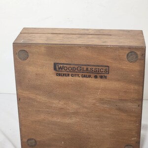 Puede incluir: Una caja rectangular de madera con acabado marr&oacute;n natural. La cara frontal presenta un logotipo negro estampado que dice WOODGLASSICS CULVER CITY, CALIF. 1974. Cuatro almohadillas circulares de fieltro est&aacute;n adheridas a las esquinas de la base.