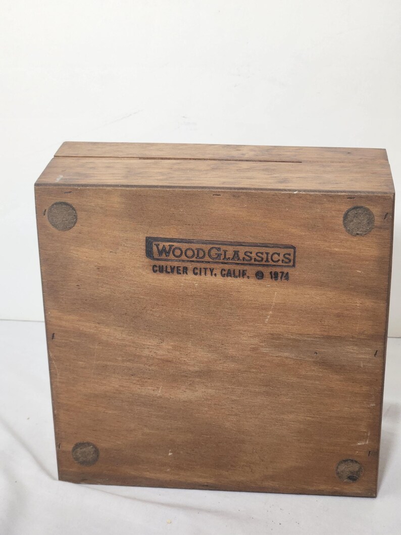 Puede incluir: Una caja rectangular de madera con acabado marr&oacute;n natural. La cara frontal presenta un logotipo negro estampado que dice WOODGLASSICS CULVER CITY, CALIF. 1974. Cuatro almohadillas circulares de fieltro est&aacute;n adheridas a las esquinas de la base.