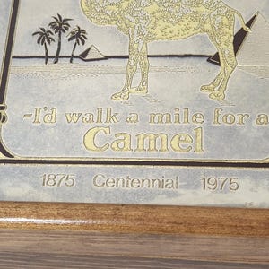 Puede incluir: Una placa rectangular con una ilustraci&oacute;n dorada de un camello, palmeras y pir&aacute;mides sobre un fondo claro. El texto dice: -I'd walk a mile for a Camel, 1875 Centennial 1975. La placa est&aacute; montada en un marco de madera sencillo.