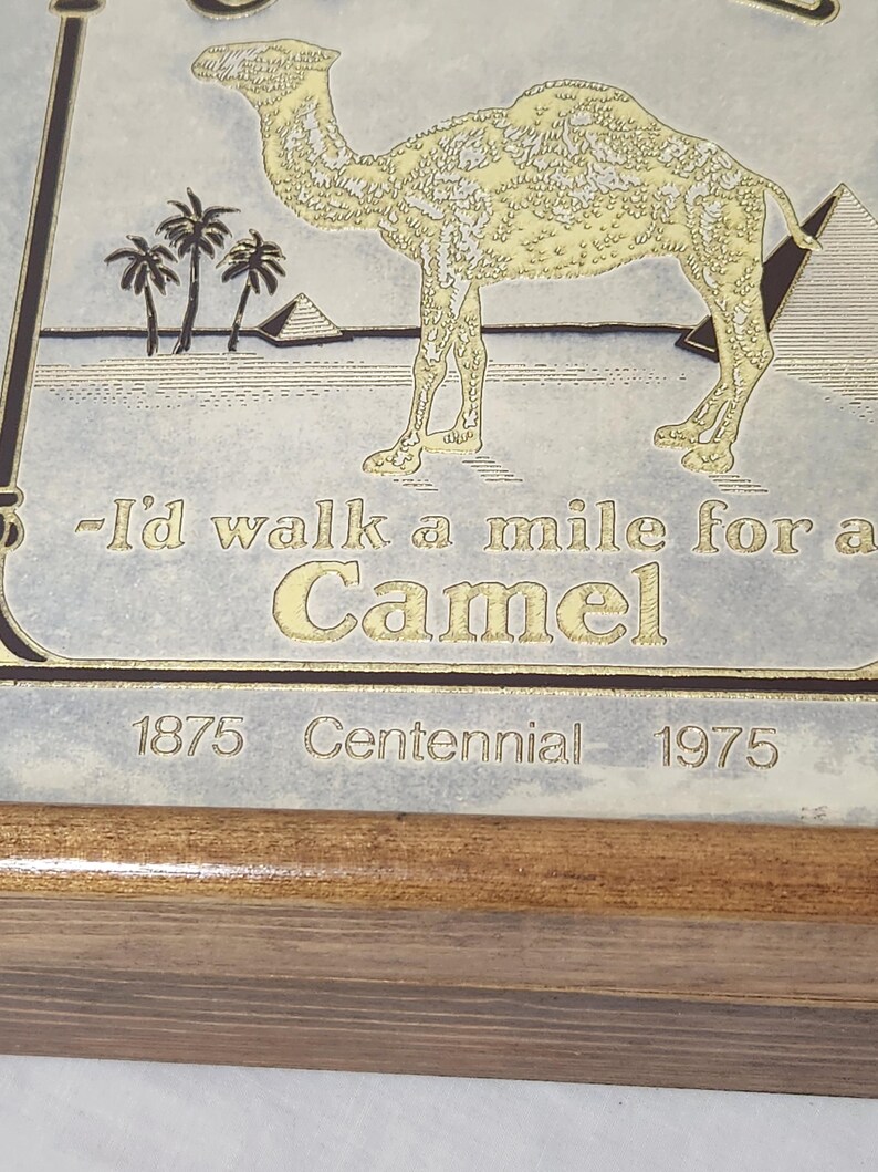 Puede incluir: Una placa rectangular con una ilustraci&oacute;n dorada de un camello, palmeras y pir&aacute;mides sobre un fondo claro. El texto dice: -I'd walk a mile for a Camel, 1875 Centennial 1975. La placa est&aacute; montada en un marco de madera sencillo.
