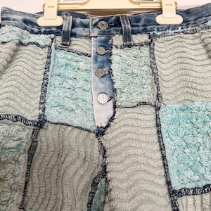 May include: A pair of blue denim shorts with a patchwork design. The shorts are made of different textures of blue fabric, including a fuzzy, textured fabric. The shorts have a button fly and a high waist.