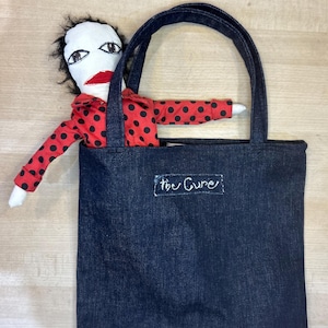 May include: A dark blue denim tote bag with black handles. A handmade doll with a white face, black hair, and a red polka dot shirt is partially visible. The text "the Cure" is written on the bag.