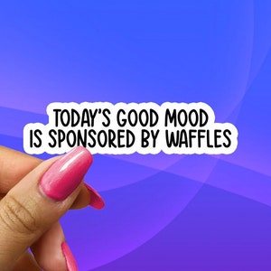 May include: A white sticker with the text "TODAY'S GOOD MOOD IS SPONSORED BY WAFFLES" in black, held by a hand with pink nail polish against a blue and purple gradient background.