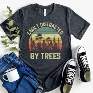 May include: A dark grey t-shirt with a vintage-style graphic that says "Easily Distracted By Trees". The graphic features a sunset with a forest silhouette.