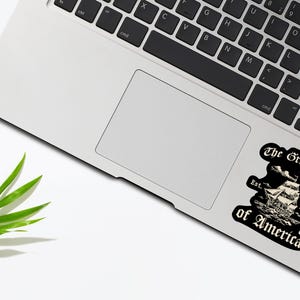 May include: A black and white sticker with a vintage illustration of a ship sailing on the water. The text "The Great Gulf of America" is written in a gothic font with the year "2025" and the word "Est." below the ship.
