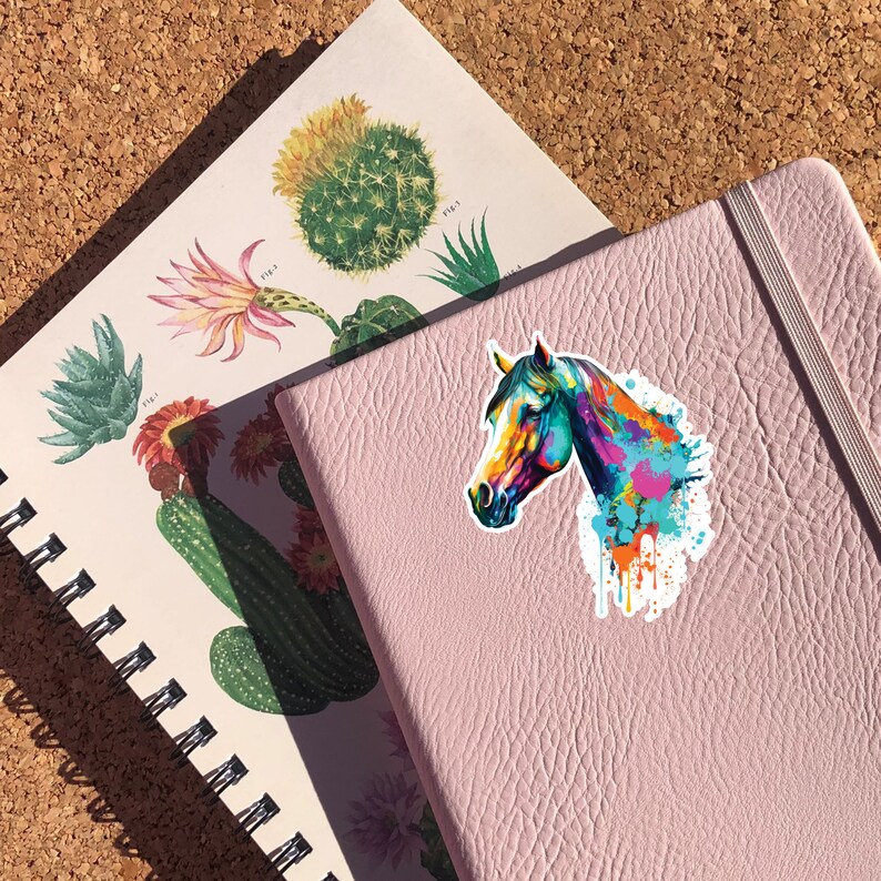 May include: A colorful horse head sticker with a watercolor effect. The horse is painted in shades of blue, green, orange, and pink. The sticker is on a pink notebook.