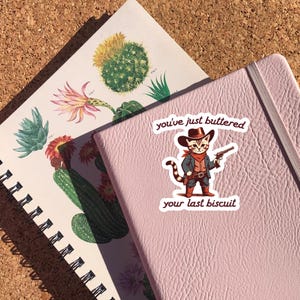 May include: A pink notebook with a sticker of a cat wearing a cowboy hat and holding a gun. The sticker says "You've just buttered your last biscuit."