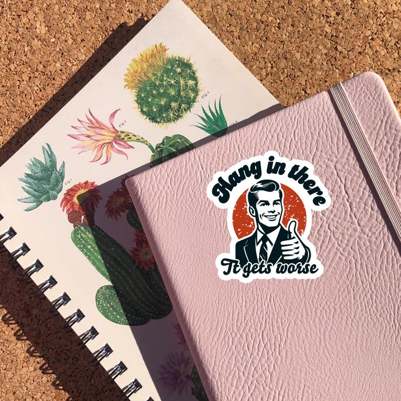 May include: A pink notebook with a sticker that says "Hang in there It gets worse" with a retro illustration of a man giving a thumbs up.