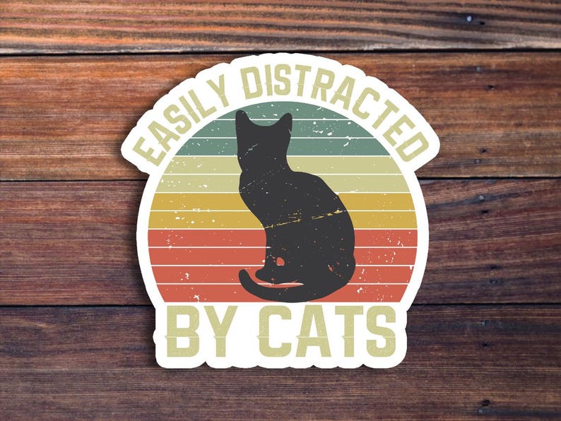 May include: A vintage-style sticker with a black silhouette of a cat sitting in front of a sunset. The sticker says "Easily Distracted By Cats".